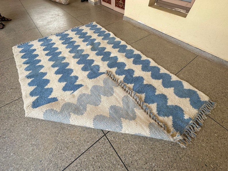 Pu&ograve; includere: Un tappeto rettangolare con sfondo color crema e un motivo ripetuto di zigzag blu. Il tappeto ha un bordo con frange ed &egrave; parzialmente arrotolato, rivelando il retro.