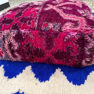 Pu&ograve; includere: Un pouf quadrato e morbido nei toni del rosa e del viola, con un design strutturato e fantasia. Il pouf &egrave; posizionato su un tappeto blu e bianco a zig-zag, creando un contrasto vibrante. Questo pezzo decorativo aggiunge colore e comfort.