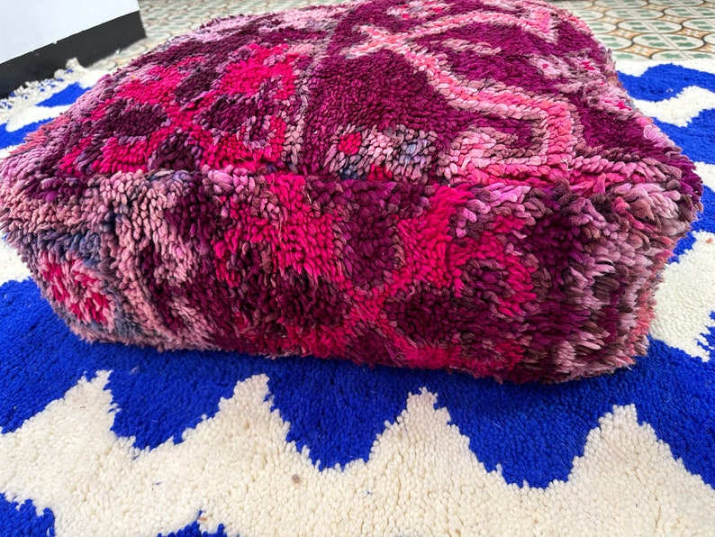 Pu&ograve; includere: Un pouf quadrato e morbido nei toni del rosa e del viola, con un design strutturato e fantasia. Il pouf &egrave; posizionato su un tappeto blu e bianco a zig-zag, creando un contrasto vibrante. Questo pezzo decorativo aggiunge colore e comfort.