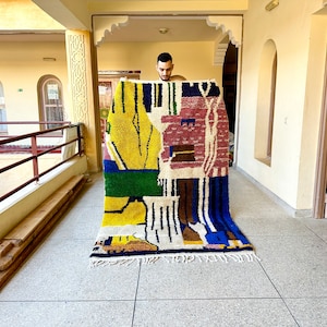 Puede incluir: Una persona sostiene una alfombra abstracta colorida con formas geom&eacute;tricas y patrones en tonos de amarillo, verde, azul, rojo y blanco. La alfombra tiene un borde con flecos.