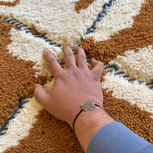 May include: A hand resting on a white and brown patterned rug with a black bracelet with a gold charm.