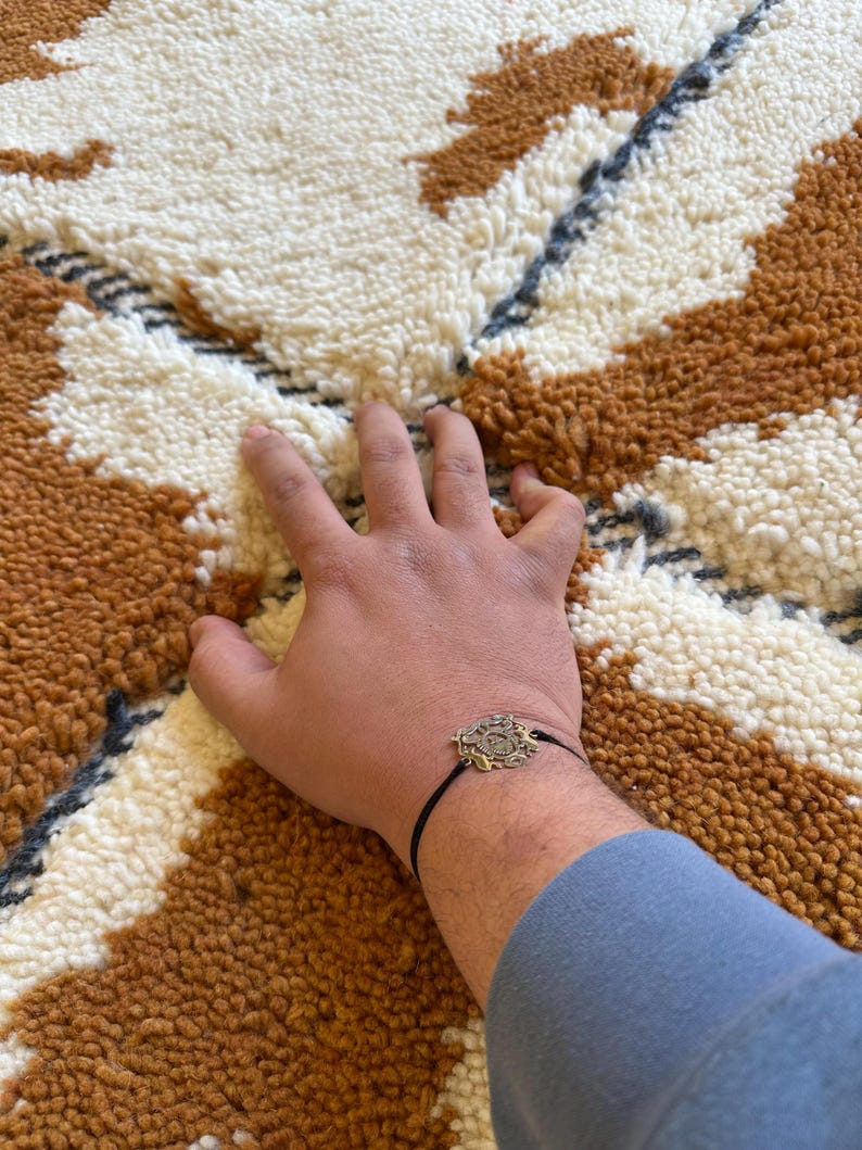 May include: A hand resting on a white and brown patterned rug with a black bracelet with a gold charm.