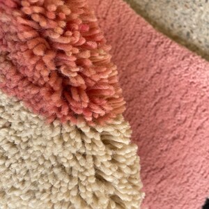 May include: Close-up of a pink, white, and black rug with a fluffy texture. The rug is made of different colored yarns woven together to create a geometric pattern.