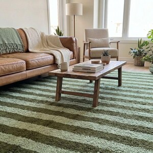 May include: A living room scene featuring a brown leather sofa with green and beige blankets, a wooden coffee table, and a striped green rug. A beige armchair and various potted plants add to the cozy atmosphere. The rug has alternating stripes of light and dark green.