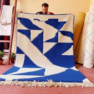 Könnte beinhalten: Ein großer, rechteckiger Teppich mit einem geometrischen Design in Blau und Creme. Der Teppich hat ein sich wiederholendes Muster aus abstrakten Formen und einen Fransenrand. Der Teppich wird in Innenräumen präsentiert, eine Person hält ihn hoch.