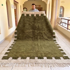 May include: A large, rectangular olive green rug with a cream border and a geometric design. The rug has a shaggy texture and a fringe along the bottom edge. The rug is being held up by a person.
