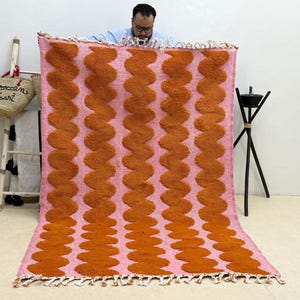 Puede incluir: Una alfombra rectangular grande con un fondo rosa y patrones ondulados naranjas repetidos. La alfombra tiene un borde con flecos. La alfombra es un revestimiento decorativo para el suelo.