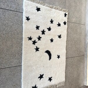 May include: A white rug with a black crescent moon and stars design. The rug has a fringe border.