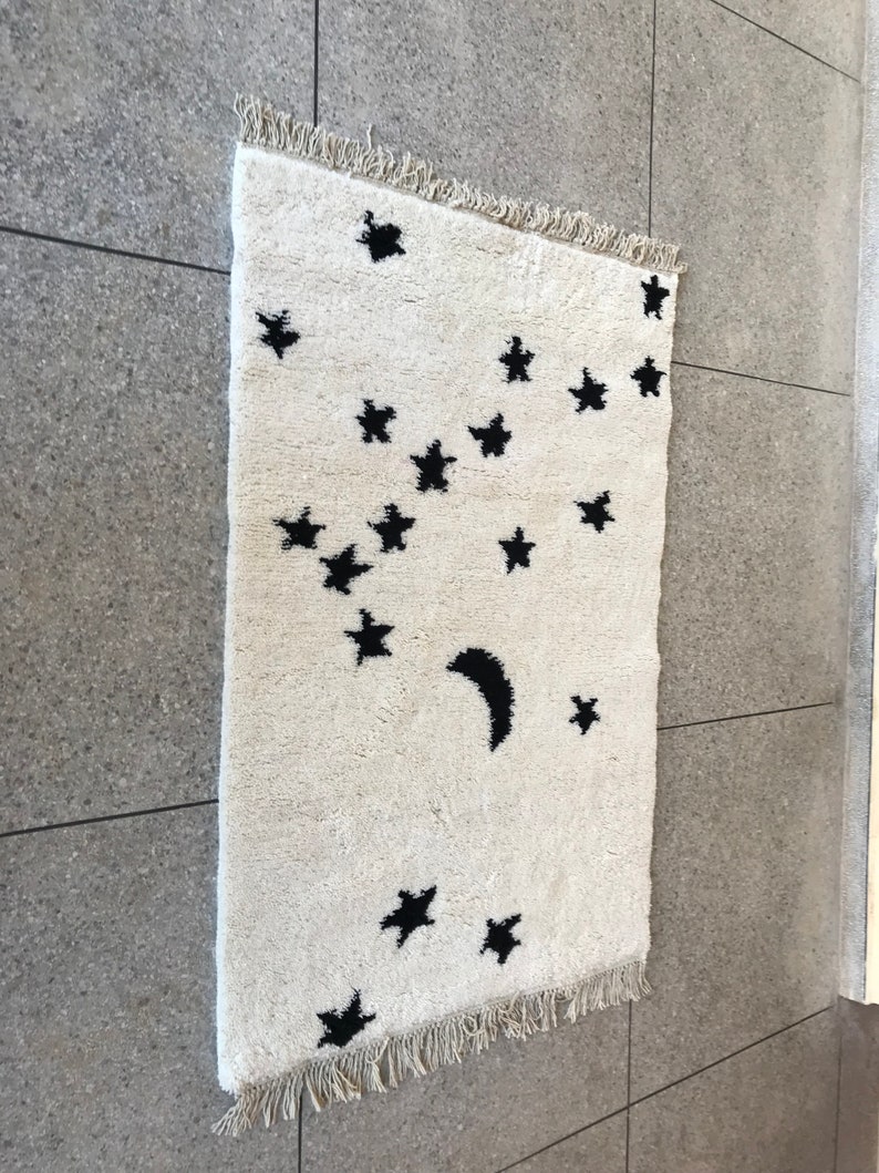 May include: A white rug with a black crescent moon and stars design. The rug has a fringe border.