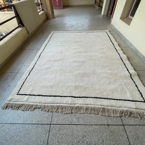 May include: A white rug with a black border and fringe. The rug is laid out on a tiled floor.