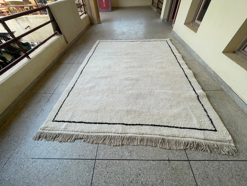 May include: A white rug with a black border and fringe. The rug is laid out on a tiled floor.