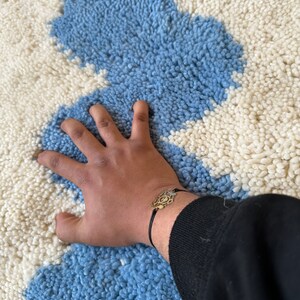 Pu&ograve; includere: Un tappeto a motivi blu e bianchi con una superficie strutturata. Un braccialetto color oro con un ciondolo decorativo &egrave; visibile su un polso.