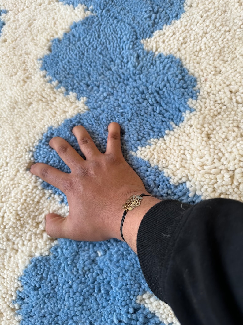 Pu&ograve; includere: Un tappeto a motivi blu e bianchi con una superficie strutturata. Un braccialetto color oro con un ciondolo decorativo &egrave; visibile su un polso.