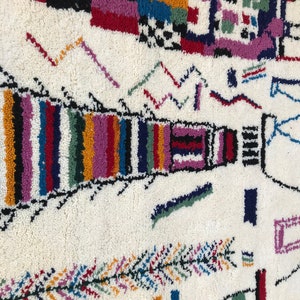 May include: A white rug with a colorful geometric pattern. The rug features a variety of shapes and colors, including red, blue, green, yellow, and purple. The rug is made of wool and has a soft, plush texture.