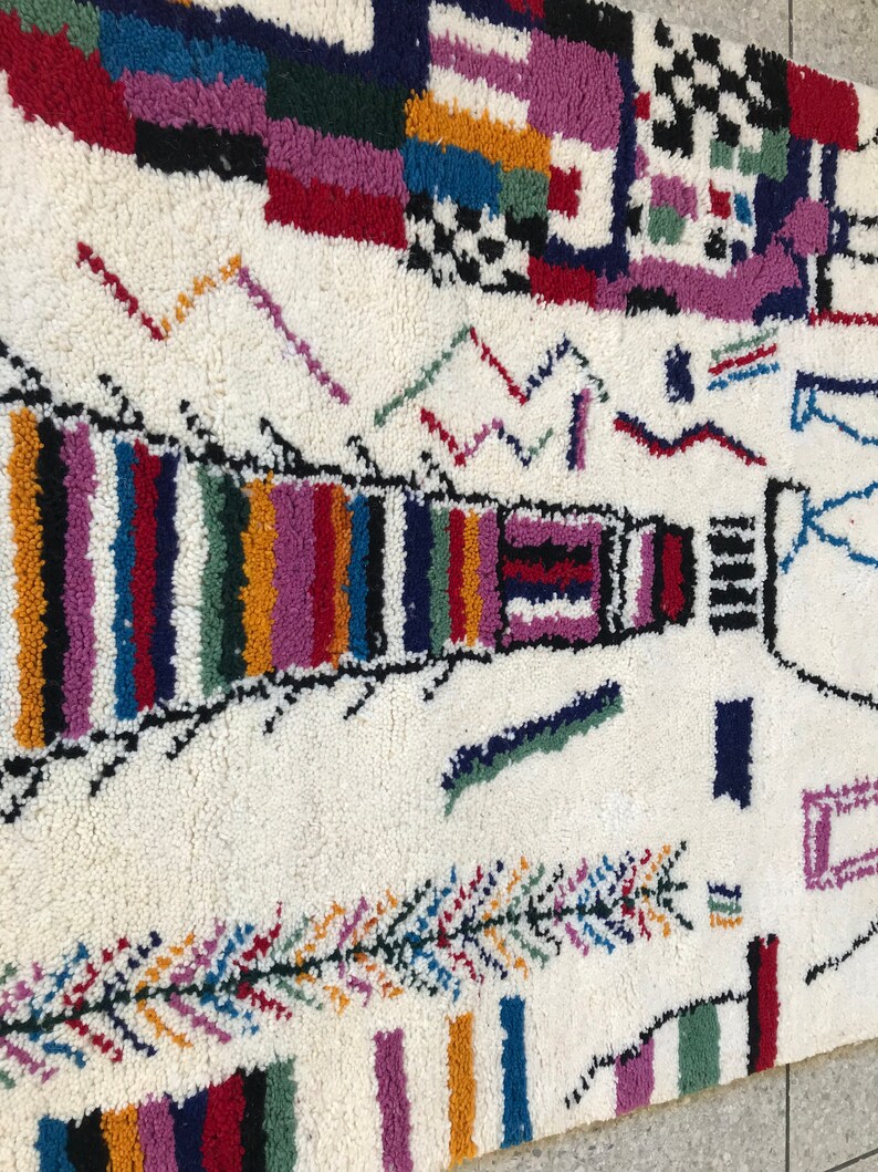 May include: A white rug with a colorful geometric pattern. The rug features a variety of shapes and colors, including red, blue, green, yellow, and purple. The rug is made of wool and has a soft, plush texture.
