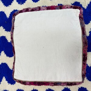 Pu&ograve; includere: Cuscino quadrato da pavimento con parte superiore bianca e bordo strutturato colorato nei toni del rosa, viola e bordeaux. Il cuscino &egrave; appoggiato su un tappeto blu e bianco a zig-zag.