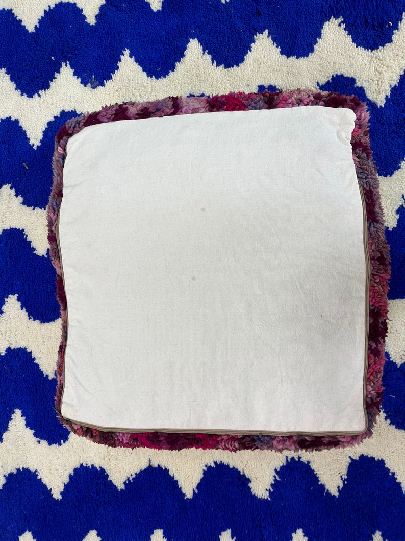 Pu&ograve; includere: Cuscino quadrato da pavimento con parte superiore bianca e bordo strutturato colorato nei toni del rosa, viola e bordeaux. Il cuscino &egrave; appoggiato su un tappeto blu e bianco a zig-zag.