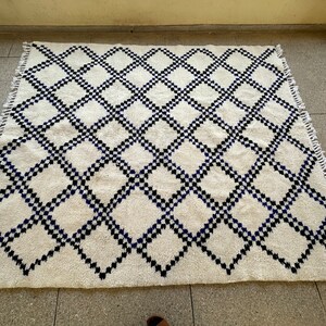 May include: A white rug with a black and blue diamond pattern. The rug has a fringe border.
