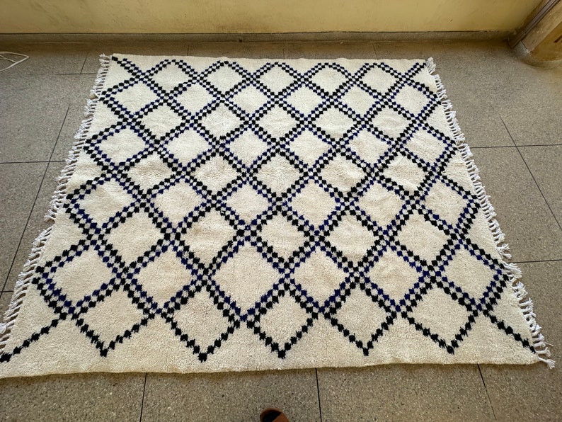 May include: A white rug with a black and blue diamond pattern. The rug has a fringe border.