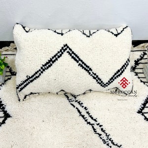 Peut inclure: Tapis marocain blanc et noir avec un motif géométrique blanc et noir. Un coussin blanc avec un motif géométrique noir est posé sur le tapis. Le tapis porte le texte "MOROCCAN" en noir.
