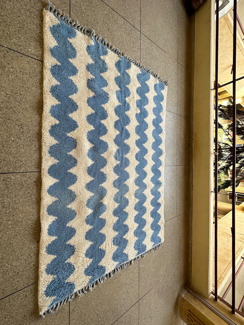 Pu&ograve; includere: Un tappeto rettangolare con un motivo ondulato ripetuto in azzurro e bianco sporco. Il tappeto ha un bordo sfrangiato ed &egrave; realizzato in un materiale strutturato. Il tappeto &egrave; esposto su un pavimento piastrellato.