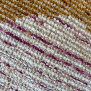 May include: Close-up of a woven fabric with a diagonal pattern. The fabric is primarily beige with thin stripes of pink and brown.