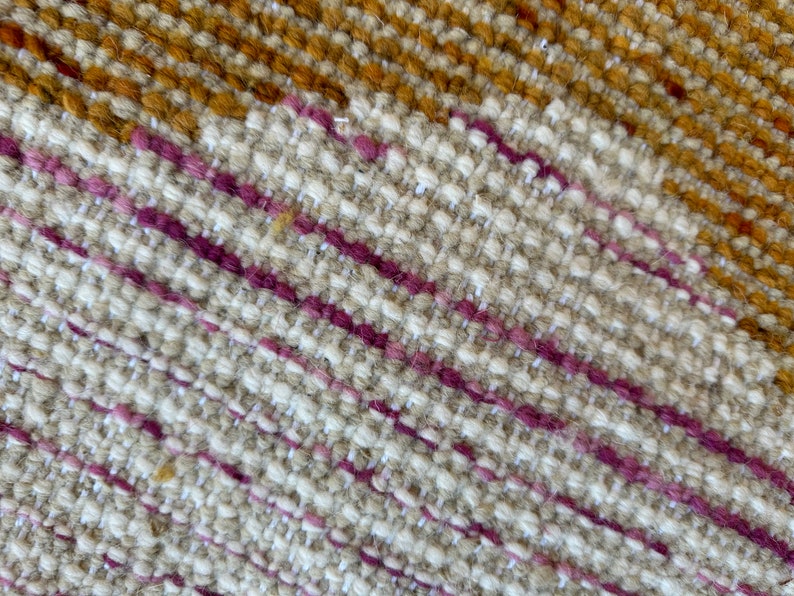 May include: Close-up of a woven fabric with a diagonal pattern. The fabric is primarily beige with thin stripes of pink and brown.