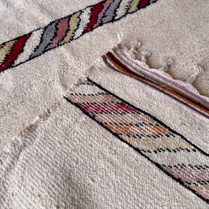 Puede incluir: Una alfombra blanca con un patr&oacute;n de rayas de colores. La alfombra tiene un borde de flecos y est&aacute; hecha de un material suave y texturizado.