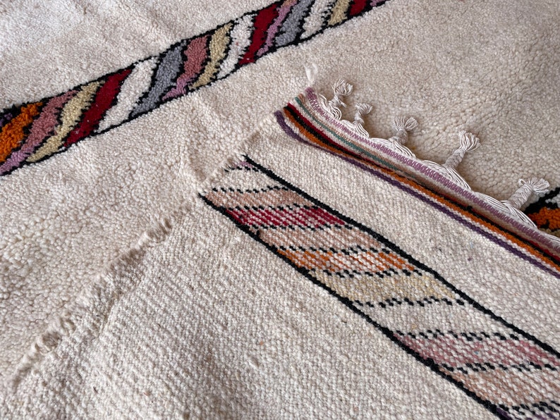 Puede incluir: Una alfombra blanca con un patr&oacute;n de rayas de colores. La alfombra tiene un borde de flecos y est&aacute; hecha de un material suave y texturizado.