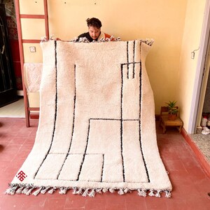 May include: A white and black geometric patterned rug with fringe. The rug is being held up by a person.