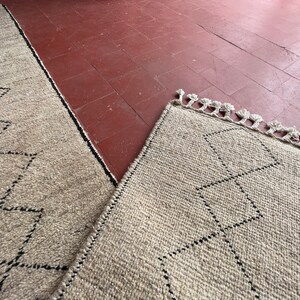 May include: Two beige rugs with a black diamond pattern and fringe edges. The rugs are laid out on a red tiled floor.
