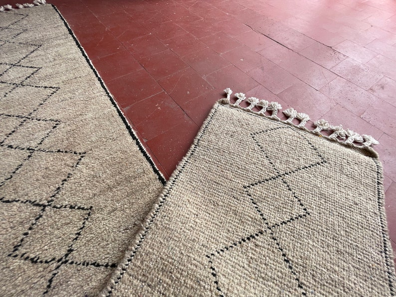 May include: Two beige rugs with a black diamond pattern and fringe edges. The rugs are laid out on a red tiled floor.