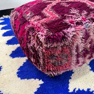 Pu&ograve; includere: Un pouf quadrato e morbido nei toni del rosa e del viola, con un design strutturato e fantasia. Il pouf &egrave; appoggiato su un tappeto blu e bianco a fantasia, creando un contrasto vibrante. Questo elemento decorativo aggiunge un tocco di colore e comfort a qualsiasi spazio abitativo.