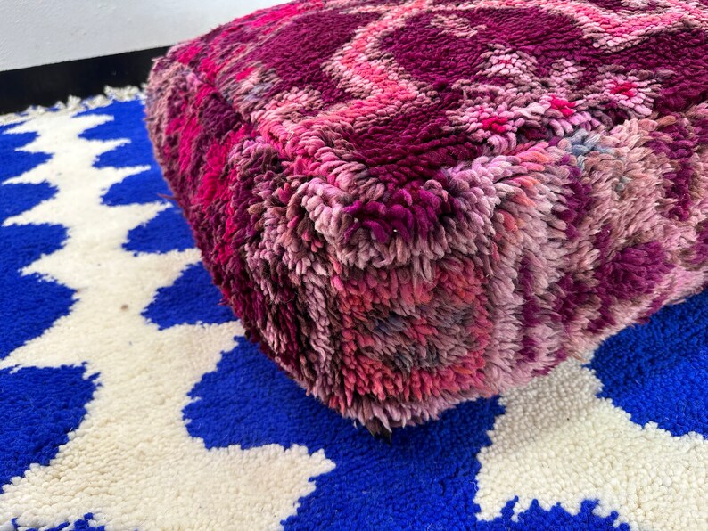 Pu&ograve; includere: Un pouf quadrato e morbido nei toni del rosa e del viola, con un design strutturato e fantasia. Il pouf &egrave; appoggiato su un tappeto blu e bianco a fantasia, creando un contrasto vibrante. Questo elemento decorativo aggiunge un tocco di colore e comfort a qualsiasi spazio abitativo.