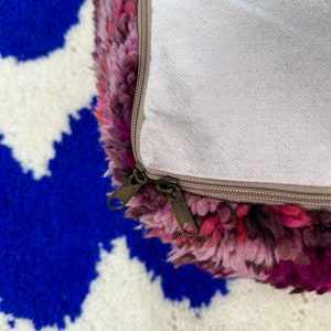 Pu&ograve; includere: Primo piano di un pouf colorato e strutturato con sfumature di rosa, viola e rosso. Il pouf ha una chiusura con cerniera ed &egrave; appoggiato su un tappeto a fantasia blu e bianco.