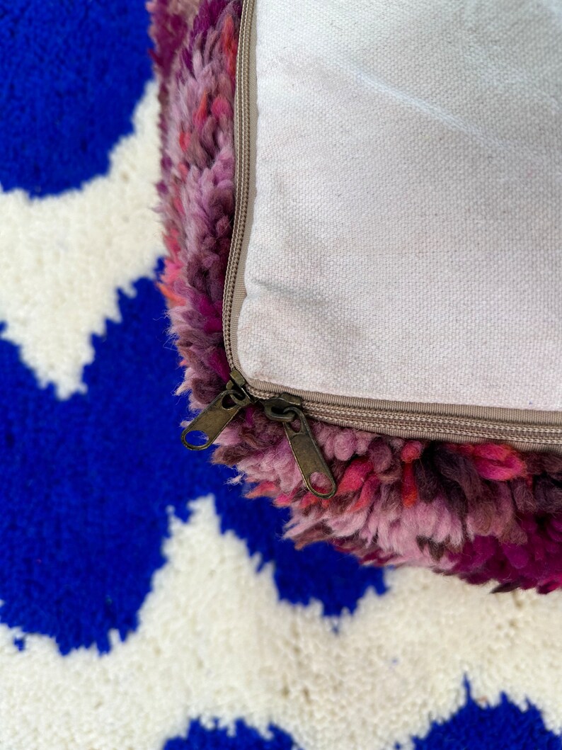 Pu&ograve; includere: Primo piano di un pouf colorato e strutturato con sfumature di rosa, viola e rosso. Il pouf ha una chiusura con cerniera ed &egrave; appoggiato su un tappeto a fantasia blu e bianco.