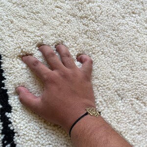 May include: A hand resting on a white fluffy rug with a black and white border. The hand is wearing a black cord bracelet with a gold charm.