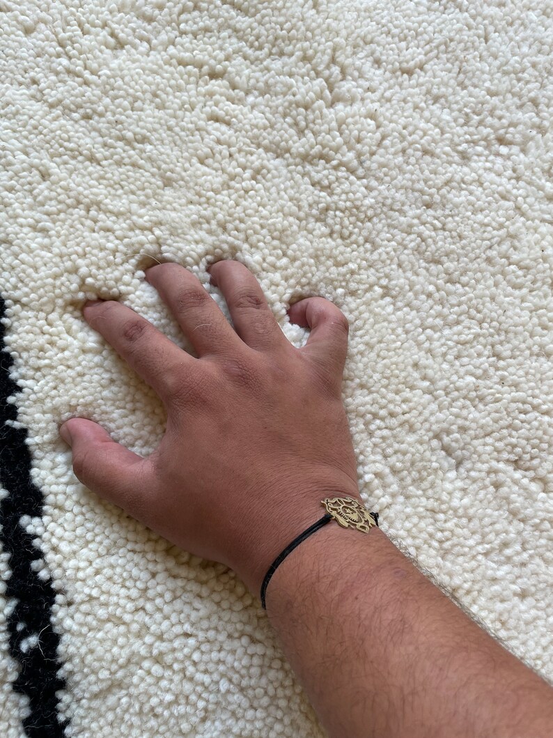 May include: A hand resting on a white fluffy rug with a black and white border. The hand is wearing a black cord bracelet with a gold charm.