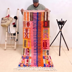 Peut inclure: Un tapis rectangulaire coloré au design vibrant. Le tapis présente une section centrale rose avec une bande verticale bleue, bordée de motifs géométriques orange, marron et autres couleurs. Le tapis a une frange blanche en bas.
