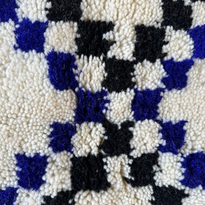 May include: Close-up of a white, black, and blue Moroccan rug with a checkered pattern. The rug is made of wool and has a soft, fluffy texture.
