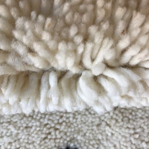 May include: Close-up of a white, fluffy rug with a long, shaggy pile. The rug has a two-toned texture, with a denser, shorter pile in the lower portion.
