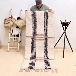 May include: Cream-colored rectangular rug with vertical black stripes and horizontal pink stripes. The rug has a fringed edge and is being held up by a person. The rug is part of a home decor collection.