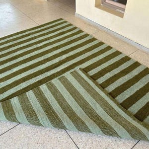May include: A green and light green striped rug. The rug is rectangular with alternating wide stripes of olive green and a lighter, muted green. The rug is partially folded, revealing the underside with the same striped pattern.