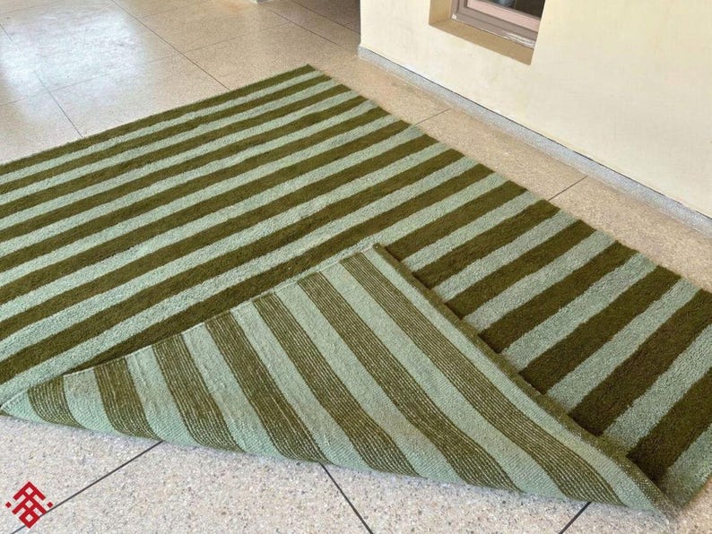 May include: A green and light green striped rug. The rug is rectangular with alternating wide stripes of olive green and a lighter, muted green. The rug is partially folded, revealing the underside with the same striped pattern.