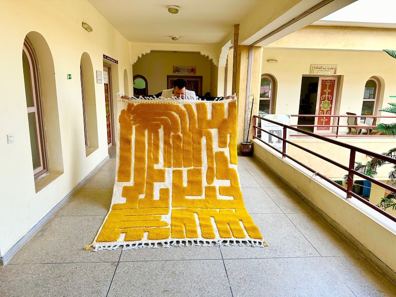 May include: A rectangular, handmade rug with a bold, abstract design in yellow and white. The rug features a geometric pattern with thick, vertical and horizontal lines. The rug has a white fringe along the bottom edge. The rug is being held up by a person.