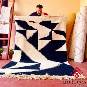 May include: A black and white geometric patterned rug with fringe. The rug is being held up by a person.