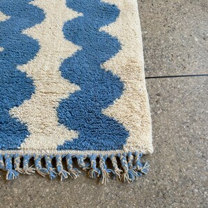 Pu&ograve; includere: Un tappeto blu e crema con un motivo a onde. Il tappeto ha una superficie strutturata e morbida e una frangia sul bordo. Il design del tappeto presenta bande alternate blu e crema, creando un effetto a zig-zag. Misure in centimetri.
