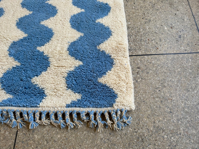 Pu&ograve; includere: Un tappeto blu e crema con un motivo a onde. Il tappeto ha una superficie strutturata e morbida e una frangia sul bordo. Il design del tappeto presenta bande alternate blu e crema, creando un effetto a zig-zag. Misure in centimetri.