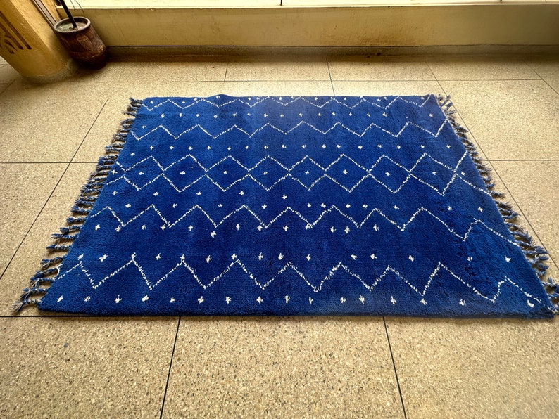 Puede incluir: Una alfombra azul y blanca con un borde de flecos. La alfombra tiene un dise&ntilde;o geom&eacute;trico con diamantes blancos y zigzags sobre un fondo azul.