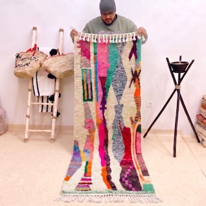 May include: A long, rectangular wool rug featuring a cream-coloured base with a vibrant, multi-coloured abstract pattern in shades of hot pink, teal, orange, purple, and grey. The rug is finished with white knotted fringe along both short ends.